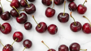 Close-up overhead shot of fresh dark red cherries scattered on a white marble countertop with soft natural window light, some with stems, showing glossy wet surfaces