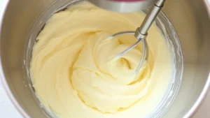Freshly whipped pale butter and sugar mixture in a stainless steel mixing bowl with electric mixer beaters showing light, fluffy texture with visible air bubbles
