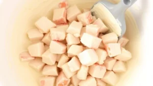 Fresh lump crab meat being gently folded into creamy mayonnaise sauce in a white mixing bowl with a rubber spatula, showing the delicate texture of the crab chunks