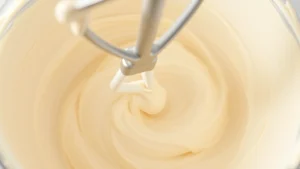 Close-up of fluffy buttercream frosting in a mixing bowl with an electric mixer beater, showing light and airy texture, soft butter-colored frosting swirled with mixer marks