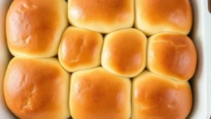 Overhead shot of soft, golden-brown Hawaiian rolls fresh from oven, steam rising, arranged in white baking pan, showing fluffy texture and glossy butter finish