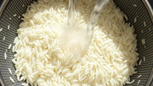 Close-up overhead shot of uncooked jasmine rice grains being rinsed in a fine-mesh strainer under flowing cool water, showing clear water running through, bright natural lighting
