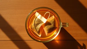 Overhead shot of two herbal tea bags steeping in a clear glass mug filled with golden-amber hot water, steam rising, natural sunlight streaming across wooden table surface, minimalist composition