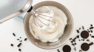 Close-up overhead view of electric mixer whipping heavy cream into fluffy white peaks in a stainless steel bowl, with crushed Oreo cookie pieces scattered nearby on a white kitchen counter
