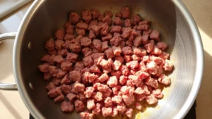 Perfectly browned ground beef in a large stainless steel skillet, meat broken into small pieces, golden-brown color, visible fond on pan bottom, natural kitchen lighting from above