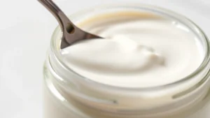Close-up of fresh creamy yogurt in glass jar with smooth texture, spoon resting on surface showing consistency, natural lighting highlighting the white color and glossy finish, minimalist white background