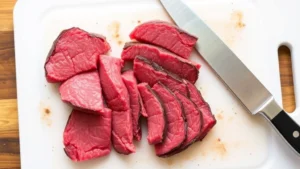 Sliced flank steak against the grain, raw beef strips on white cutting board with kitchen knife, clearly separated pieces showing proper thickness