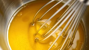 Close-up of champagne vinaigrette being whisked in a stainless steel bowl, oil and vinegar combining into creamy emulsion with visible mustard swirl, golden liquid with air bubbles, professional kitchen lighting