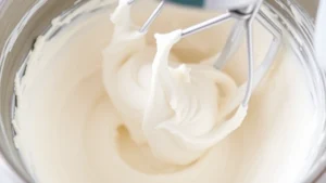 Close-up of freshly whipped ermine frosting in a stainless steel mixing bowl with electric mixer beaters visible, showing the light fluffy texture with peaks, natural kitchen lighting, cream and white tones